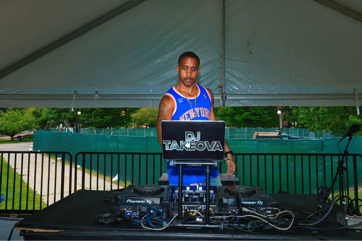 DJ Takeova behind Pioneer decks under an outdoor stage tent