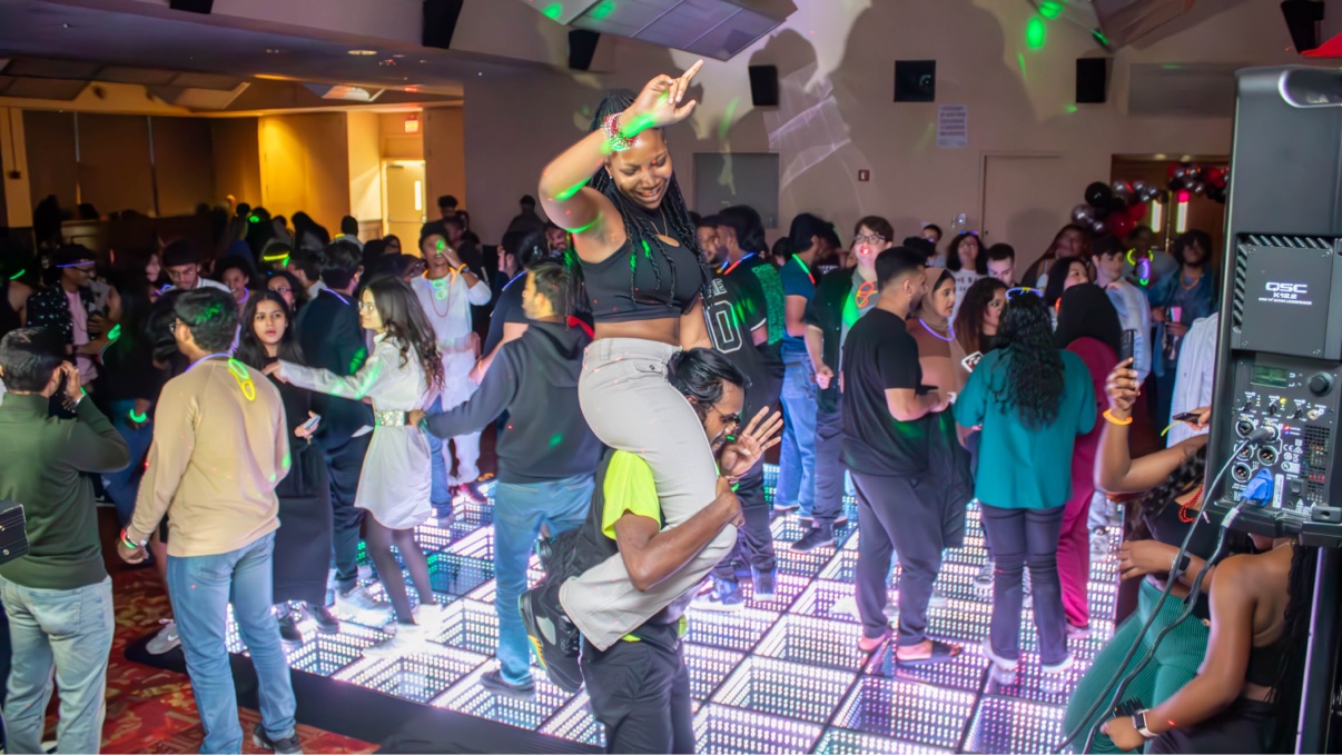 A smiling student riding on a friend's shoulders above a packed crowd on a glowing LED dance floor, one arm raised and pointing up