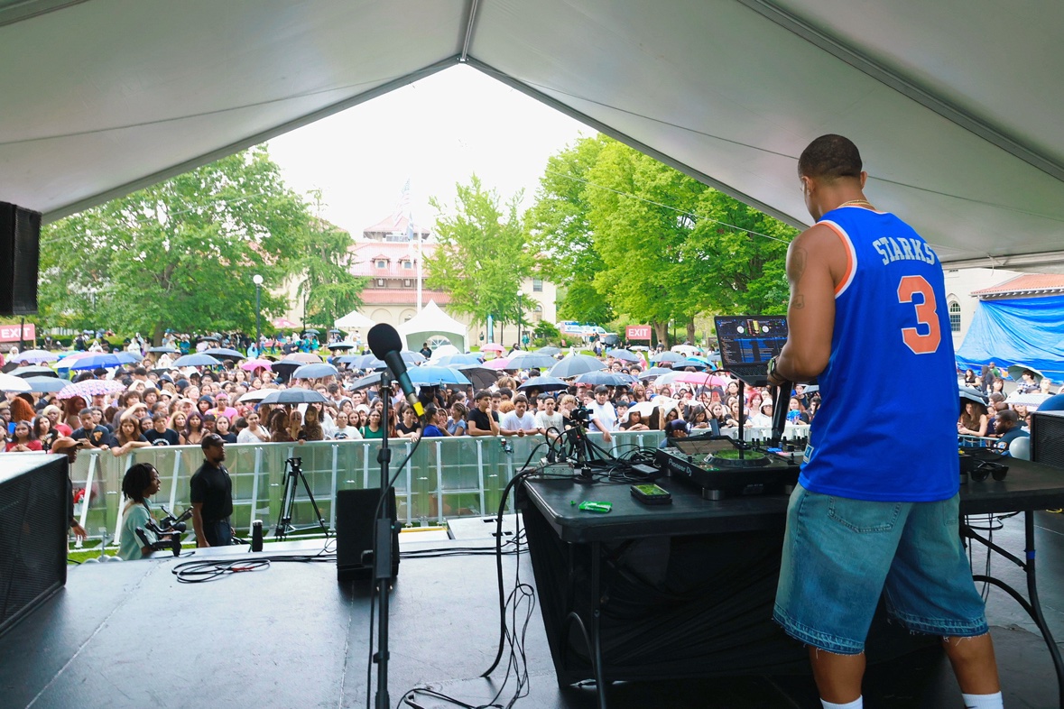 DJ Takeova in pink at the decks before a rainy outdoor crowd with umbrellas