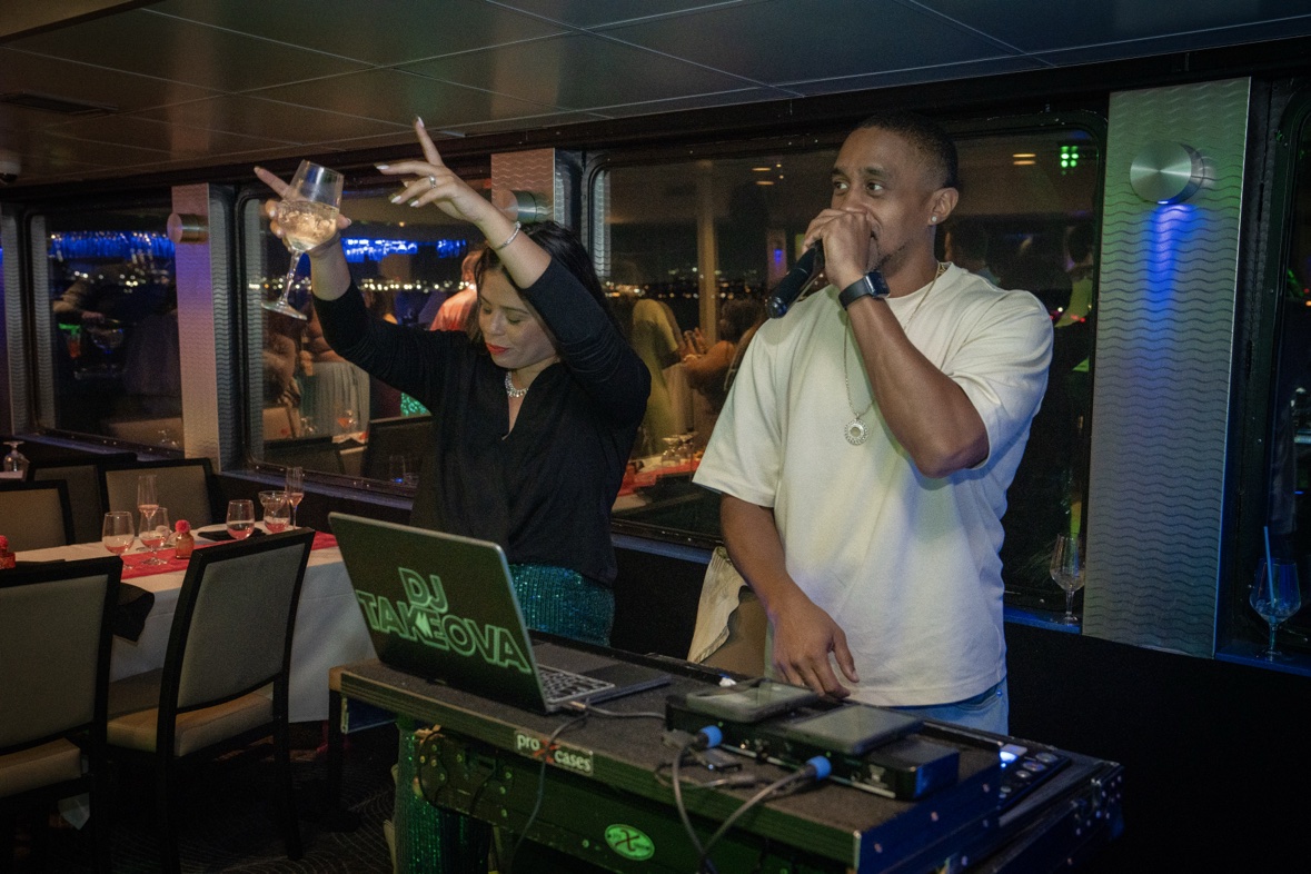 Dj Takeova on the mic at the decks during a private boat party as a guest dances with champagne